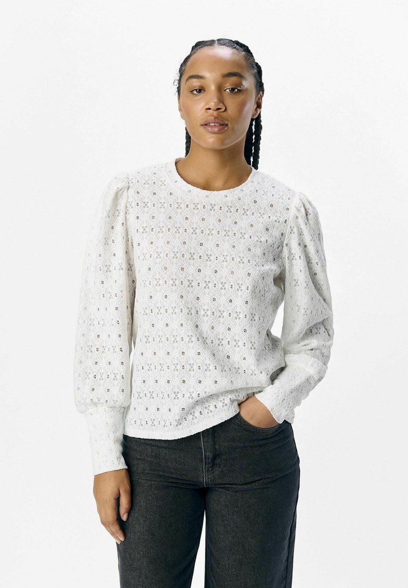 Pull en dentelle blanche avec découpes florales, tissu texturé, col rond, et longues manches bouffantes, associé à un pantalon en denim foncé.