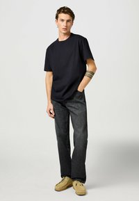 Sort kortærmet t-shirt parret med mørke denim jeans. Modellen har beige mokkasinstil sko på og har en flerstribet tatovering på håndleddet.