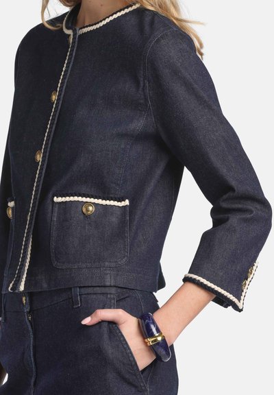 Denimjakke i mørkeblå med cremefarvet kant, guldknapper og to forlommer. Har et kort design og trekvart lange ærmer.