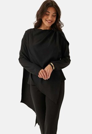 Kvinna som bär en svart asymmetrisk långärmad topp med draperad rygg, tillsammans med åtsittande svarta byxor, ler med händerna lätt sammanflätade.