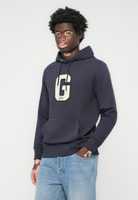 Marineblå hoodie fremstillet af bomuldsblanding, med et stort kremfarvet G-logo på brystet, justerbare snore og en frontkænguru-lomme.
