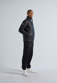 Zwarte pufferjack met horizontaal doorgestikt patroon, blauwe rits en hoge kraag. Draagt met zwarte broek en witte sneakers.