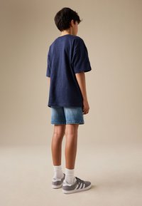 Chłopiec stoi tyłem do kamery, ubrany w luźny granatowy t-shirt, wyblakłe krótkie dżinsy, białe skarpetki i szare buty sportowe Adidas.