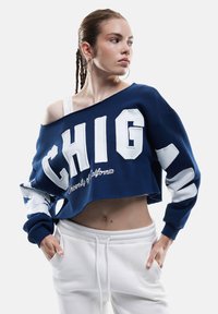 Sudadera corta azul marino con letras blancas en bloque "CHIG" y "Universidad de California", combinada con pantalones de chándal blancos.
