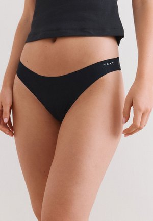 Femme portant un sous-vêtement style bikini noir avec le logo "NEXT" sur la ceinture, associé à un top court noir, sur un fond uni.