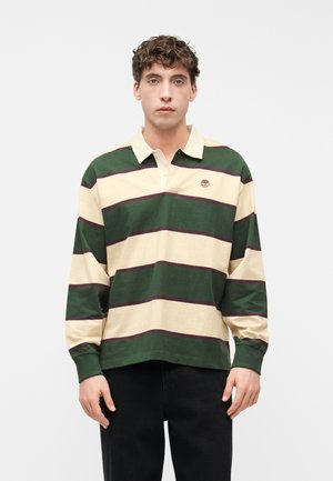 Fiatal férfi zöld és bézs csíkos hosszú ujjú rögbiingben, sötét nadrágban, egyszínű világos háttér előtt áll.