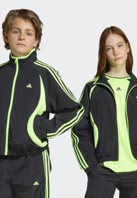 Crne i neon zelene Adidas trenirke imaju zatvaranje na patentni zatvarač, visoki ovratnik i bočne džepove, s prugastim detaljima duž rukava.