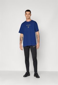 Mand iført en blå Spurs t-shirt, sorte Nike sweatpants og sorte sneakers står mod en enkel lysegrå baggrund.
