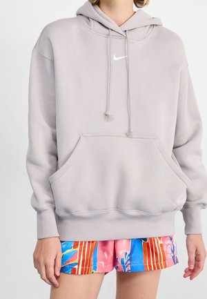 Personne portant un sweat à capuche Nike gris clair avec poche avant et cordons, associé à un short coloré à motifs bleus, roses, oranges et blancs.
