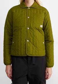 Quiltad olivgrön jacka med tryckknappar, rundad krage, två framfickor och subtil logo på vänster sida. Slät textur.