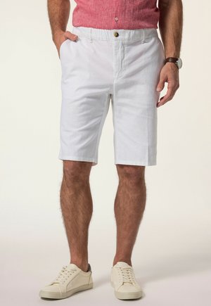 Mann trägt weiße knielange Shorts, pinkes Kurzarmhemd, weiße Sneakers und eine Armbanduhr, eine Hand in der Tasche.
