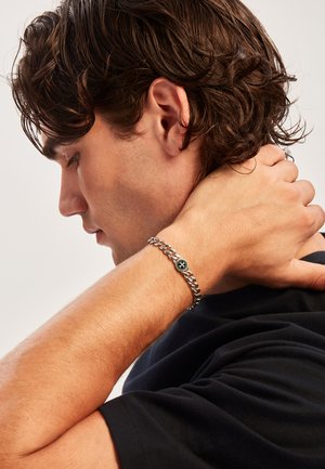 Bracciale in argento con un emblema nero ovale che presenta un design a stella, indossato su un polso contro una camicia scura.