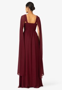 Abito bordeaux con schiena quadrata, corpino arricciato, maniche lunghe e fluenti, e tessuto texturizzato, che scende fino al pavimento in una silhouette rilassata.
