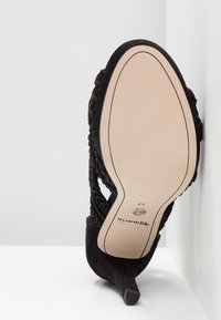 Black high-heeled shoe with a textured suede upper, cut-out design, and a pointed toe. Features a light-colored sole and a slim heel.