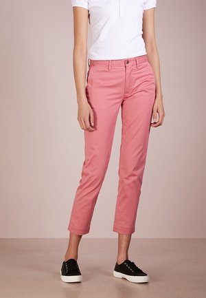 Persoon draagt roze cropped broek, een wit T-shirt met korte mouwen en zwarte sneakers, staand tegen een effen achtergrond.