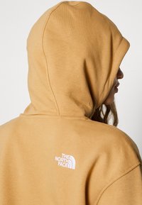 Personne portant un sweat à capuche beige avec le logo The North Face brodé en blanc sur le haut du dos, tournée vers la droite, sur fond uni.