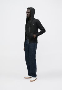 Sudadera negra con cremallera y capucha, usada sobre una camiseta negra, combinada con vaqueros azul oscuro y zapatillas blancas con suelas marrones.