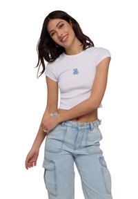 T-shirt bianca cropped con una grafica di un orso blu sul petto, maniche corte e vestibilità aderente, abbinata a pantaloni cargo blu chiaro.