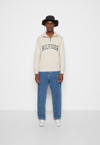 Felpa beige con zip e testo "HILFIGER", abbinata a jeans blu larghi e scarpe da ginnastica bianche. Include un cappello a secchiello nero.