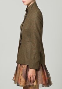 Blazer de tweed marrón con un diseño estructurado, que presenta un cuello alto y bolsillos laterales, llevado sobre una falda ligera, colorida y con capas.