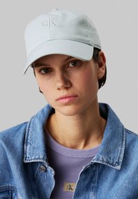Casca de un bleu deschis cu logo-ul „CK” brodat. Combinată cu o jachetă din denim și o cămașă cu nervuri violetă, care are un eticheta cu logo. Fundal neutru.