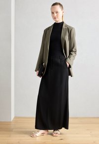 Blazer vert olive sur un col roulé noir, jupe maxi noire et sandales beiges avec des nœuds. Tient une pochette noire. Textures lisses.