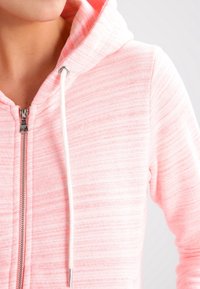 Sudadera con cremallera de color rosa claro con una textura suave, patrón a rayas y cordones blancos. Herrajes de cremallera plateados visibles en el cuello.