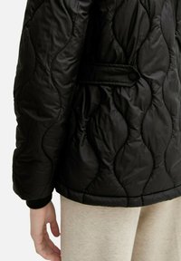 Veste matelassée noire avec un motif ondulé texturé, dotée d'un accent latéral à boutons et de poignets côtelés. Portée avec un pantalon gris clair.