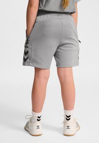 Kind draagt grijze sportbroek met ritssluitingen, witte sokken en witte sneakers met zwarte hummel-logo's, staand tegen een witte achtergrond.