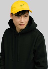 Żółta bawełniana czapka z haftowanym, kolorowym wzorem ptaka, noszona z czarną bluzą z kapturem. Prosty, swobodny styl.