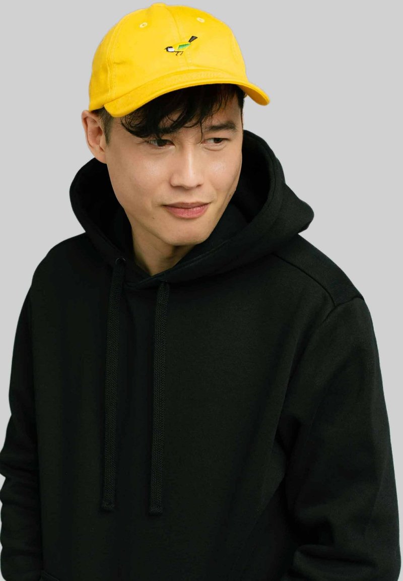 Żółta bawełniana czapka z haftowanym, kolorowym wzorem ptaka, noszona z czarną bluzą z kapturem. Prosty, swobodny styl.