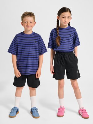 To børn står side om side iført blå og sortstribede skjorter, sorte shorts, hvide sokker og farverige sneakers.