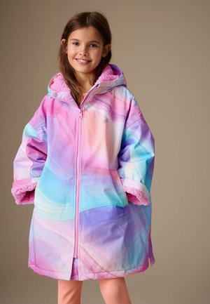 Ung pige smiler, iført en pastel regnbuefarvet oversized hættetrøje med pink fleecefor og ribstrikkede orange leggings.