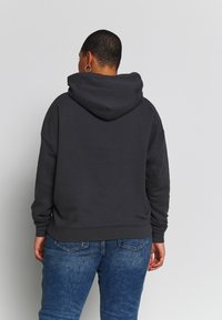 Personne aux cheveux courts portant un sweat à capuche noir et un jean bleu, vue de dos sur un fond uni gris clair.