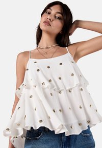 Weißes ärmelloses Top mit geschichteten Rüschen, verziert mit goldenen Pailletten. Der Stoff hat ein leichtes, strukturiertes Finish. Kombiniert mit Denim-Shorts.