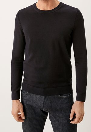 Mann trägt einen schlichten schwarzen Pullover mit Rundhalsausschnitt und lange Ärmel sowie dunkelgraue Jeans, steht vor einem hellen neutralen Hintergrund.