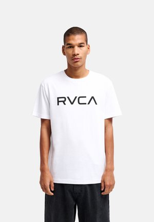 Junger Mann mit kurzen Haaren, der ein weißes T-Shirt mit schwarzem "RVCA"-Logo und schwarze Hosen trägt, steht vor einem schlichten, hellen Hintergrund.