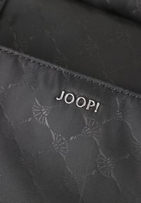 Tissu noir texturé avec un motif subtil, arborant le logo argenté « JOOP ! ». La surface présente une finition lisse et des bords cousus.