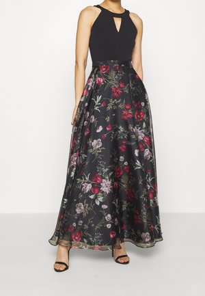 Woman wearing sleeveless black top with keyhole neckline and long black floral skirt with red and pink flowers, paired with black heels.