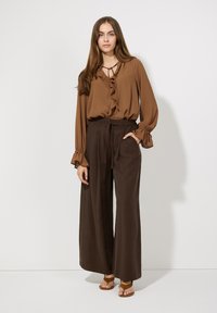 Braun rüschige Bluse mit einem Bindekragen, kombiniert mit hochgeschnittenen, weit geschnittenen braunen Hosen. Sandalen vervollständigen das Outfit.