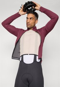 Fahrradbekleidung mit einem weinroten Langarmtrikot mit Reißverschluss, einer beigen strukturierten Basisschicht und schwarzen gepolsterten Radsportshorts.