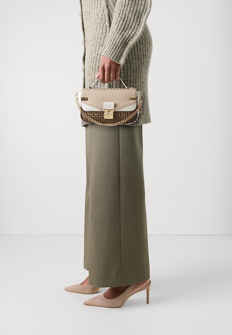 Sac à main beige à motifs avec quincaillerie dorée, bandoulière en chaîne et accents blancs. Porté avec un pantalon large vert olive et des talons beiges.