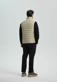 Uomo vestito con un gilet trapuntato beige su una camicia nera a maniche lunghe, pantaloni neri e scarpe da trekking marroni, in piedi con le mani in tasca, rivolto di spalle.