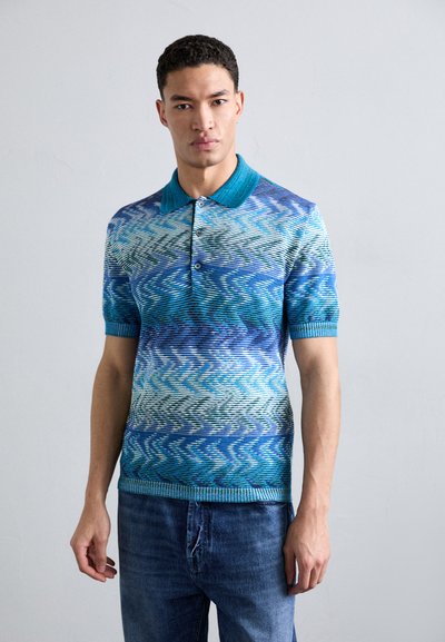 Polo a maniche corte con motivo blu e verde. Presenta un colletto turchese a tinta unita e un design a maglia testurizzato. Indossato con jeans blu.