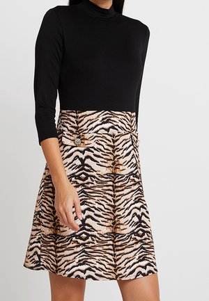 Frau trägt ein schwarzes Kleid mit Dreiviertelärmeln, einem Rock mit Tigerprint und dekorativen Knöpfen an der Taille.