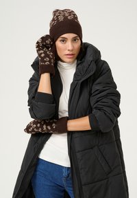 Bruine gebreide muts met luipaardmotief; bijpassende bruine wanten. Het model draagt een zwarte pufferjas en een witte coltrui, met spijkerbroek.