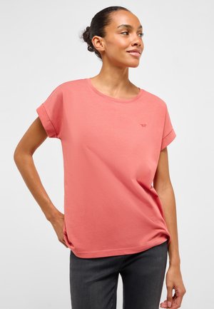 Mujer con el cabello recogido en un moño, vistiendo una camiseta coral de manga corta con puños enrollados y pantalones oscuros, de pie con una mano en la cadera, mirando hacia la derecha.