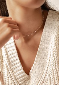 Collar de choker en oro rosa con eslabones alargados y facetados, lucido en un cuello al descubierto, combinado con un suéter de crema texturizado que presenta un cuello acanalado.