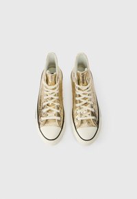 Tênis de cano alto em dourado metálico, apresentando uma superfície texturizada, biqueira de borracha branca e atacadores creme. Detalhes pretos ao longo da sola.