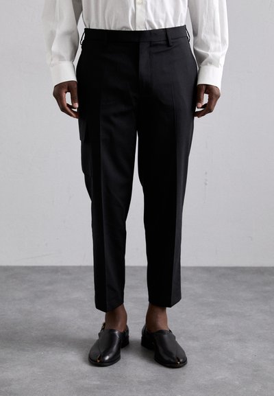 Pantaloni negri, adaptați, cu o croială slim, buzunare laterale și detalii cu pliuri, asortați cu pantofi negri slip-on pe un podea gri deschis.